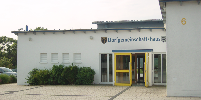 Die Eingangstüre ist gelb, über der Türe steht zwischen zwei Wappen "Dorfgemeinschaftshaus"