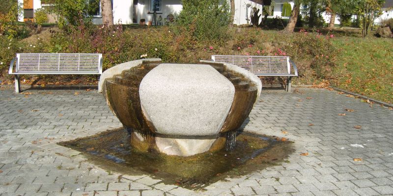  Der Brunnen steht mittig auf einem Platz, hinten sind zwei Bänke 