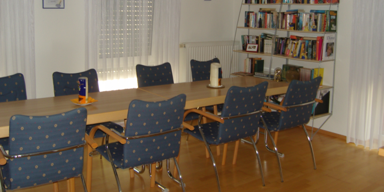 Links ist ein langer Tisch mit acht Stühlen, rechts ein Bücherregal