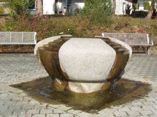 Der Brunnen steht mittig auf einem Platz, hinten sind zwei Bänke